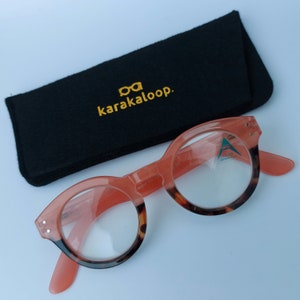 Könnte beinhalten: Eine runde Lesebrille mit rosafarbenem Rahmen und Schildpatt-Akzenten. Die Brille befindet sich in einem schwarzen Filz-Etui mit dem in Gold gestickten Text "karakaloop."