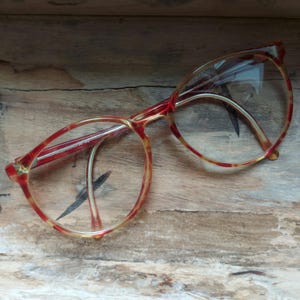 May include: Red and yellow patterned eyeglasses with clear lenses. The frames have a retro design, with a rounded shape and a marbled effect. The glasses are set against a wooden background.