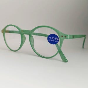 Pode incluir: Óculos de leitura redondos verdes com um rótulo de lente asférica +1.00. Os óculos têm uma armação fina e leve.
