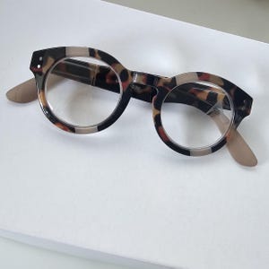 May include: Round reading glasses featuring a brown and black tortoiseshell pattern on the frames. The clear lenses have the text +2.00 printed on the right side. The temples are a solid light beige color, and the frame front has small silver dots.
