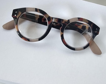 Könnte beinhalten: Runde Lesebrille mit einem braun-schwarzen Schildpattmuster auf dem Rahmen. Die klaren Gläser tragen den Aufdruck +2.00 auf der rechten Seite. Die Bügel sind in einem einfarbigen Hellbeige gehalten und die Vorderseite hat kleine silberne Punkte.