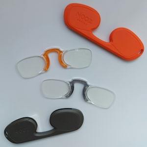 May include: An overhead shot shows three pairs of reading glasses and two cases. The glasses have clear lenses and orange or gray frames. One orange case has the text "NOOZ optics" printed on it. The cases and glasses are on a white surface.