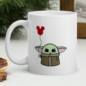 May include: White ceramic mug with a cartoon illustration of a green character holding a red balloon shaped like Mickey Mouse ears.