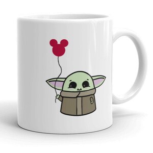 May include: White ceramic mug with a cartoon illustration of a green character holding a red balloon shaped like a Mickey Mouse head.