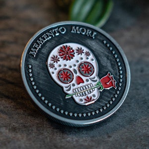 May include: A silver-toned round coin with a black background and a white sugar skull design with red accents. The skull is holding a red rose in its mouth. The text "Memento Mori" is inscribed around the edge of the coin.