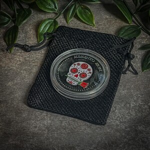 May include: A silver-colored round coin with a black velvet pouch. The coin features a white sugar skull with red flowers and a red rose. The text "Memento Mori" is inscribed around the skull.