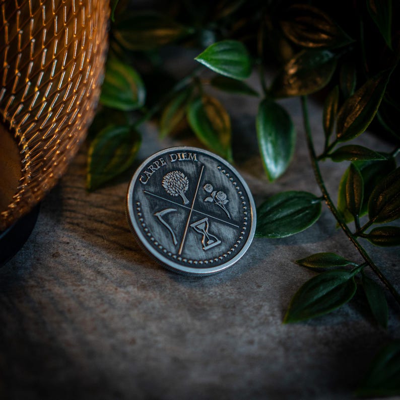 May include: A silver-toned metal pin with the words "Carpe Diem" inscribed around the edge. The pin features a tree, a rose, a scythe, and an hourglass.