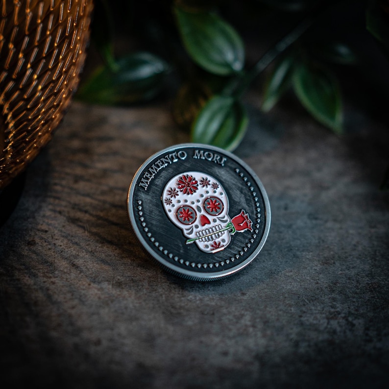 May include: A silver-toned round coin with a black background and a white sugar skull design with red accents. The skull is holding a red rose in its mouth. The text "Memento Mori" is inscribed around the edge of the coin.