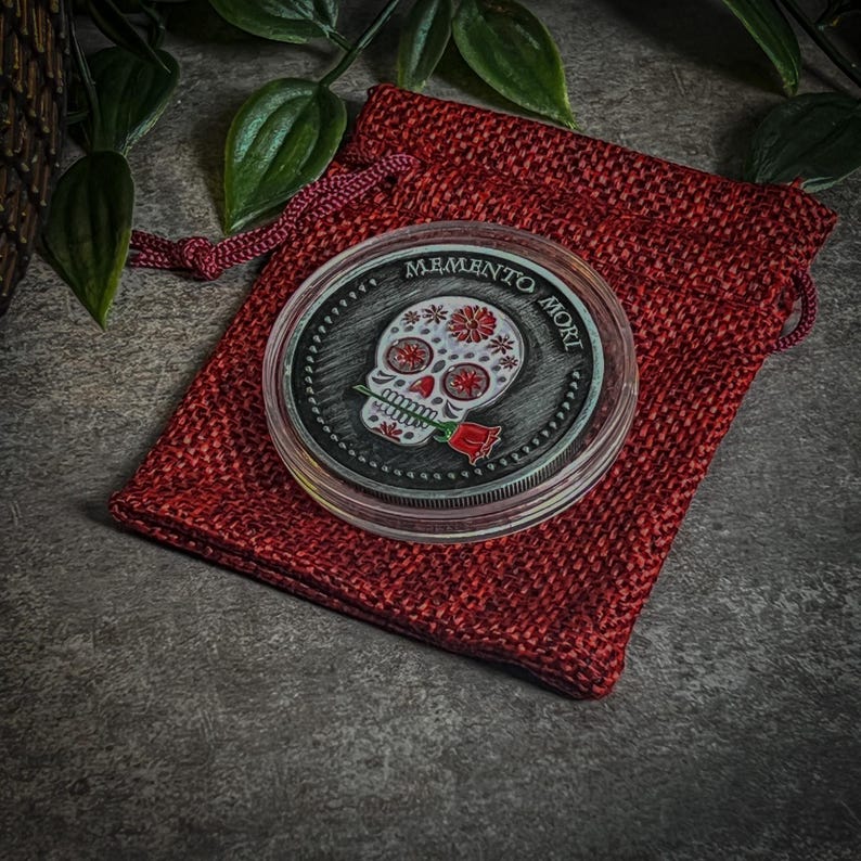 May include: A silver coin with a black and white sugar skull design and a red rose. The coin is in a red velvet pouch and the text "Memento Mori" is inscribed around the edge.