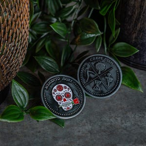 May include: Two silver-toned metal coins with black and white designs. One coin features a skull with a red rose and the text "Memento Mori". The other coin features a tree, an hourglass, a scythe, and the text "Carpe Diem".