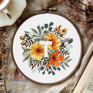May include: Cross stitch pattern featuring a floral wreath with yellow and orange flowers, green leaves, and a white letter "T" in the center. The pattern is displayed on a wooden embroidery hoop.