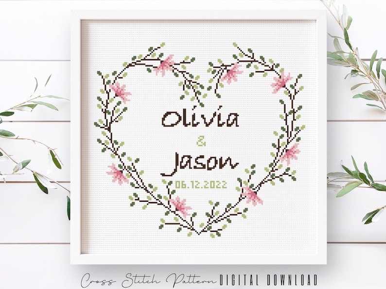 K&ouml;nnte beinhalten: Kreuzstichmuster mit einem herzf&ouml;rmigen Blumenkranz mit den Namen Olivia & Jason und dem Datum 06.12.2022.