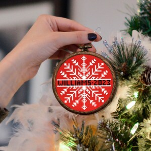 May include: A red and white snowflake bauble with the text "Williams 2023" on a wooden frame. The bauble is being held by a hand in front of a Christmas tree.