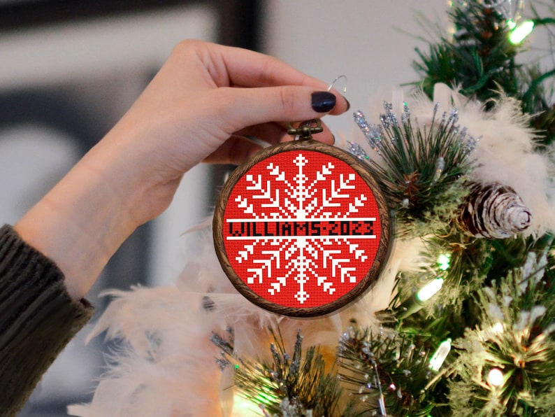 May include: A red and white snowflake bauble with the text "Williams 2023" on a wooden frame. The bauble is being held by a hand in front of a Christmas tree.