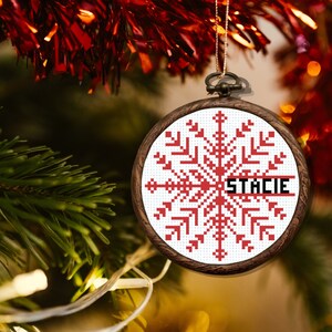 May include: A white and red cross-stitch ornament with a snowflake design and the name "Stacie" in black lettering. The ornament is hanging on a Christmas tree.