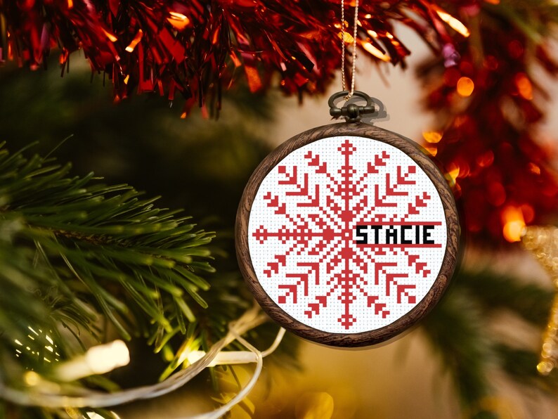 May include: A white and red cross-stitch ornament with a snowflake design and the name "Stacie" in black lettering. The ornament is hanging on a Christmas tree.
