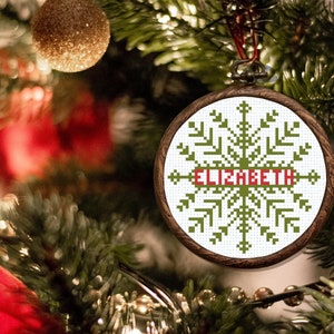 May include: A green and white snowflake bauble with the name "Elizabeth" embroidered in red on a wooden frame. The bauble is hanging on a Christmas tree.