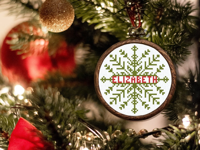 May include: A green and white snowflake bauble with the name "Elizabeth" embroidered in red on a wooden frame. The bauble is hanging on a Christmas tree.