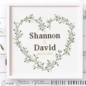 May include: Cross stitch pattern featuring a heart-shaped wreath of green leaves with the names "Shannon & David" and the date "06.25.2022" in the center.