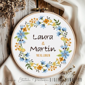 May include: Cross stitch pattern featuring a floral wreath with blue and yellow flowers, the names Laura & Martin, and the date 10.12.2025.