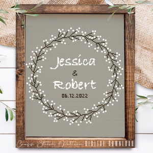 May include: Cross stitch pattern featuring a wreath of white flowers and brown branches with the text "Jessica & Robert 06.12.2022" in the center.