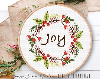 May include: Cross stitch pattern featuring a wreath of green leaves, red berries, and pink flowers with the word "Joy" in the centre.