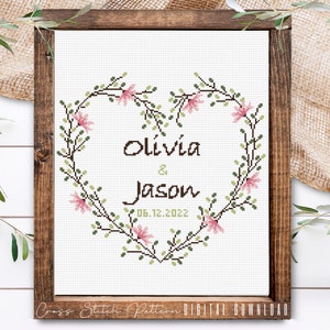 K&ouml;nnte beinhalten: Kreuzstichmuster mit einem herzf&ouml;rmigen Kranz aus gr&uuml;nen Bl&auml;ttern und rosa Bl&uuml;ten mit den Namen Olivia & Jason und dem Datum 06.12.2022.