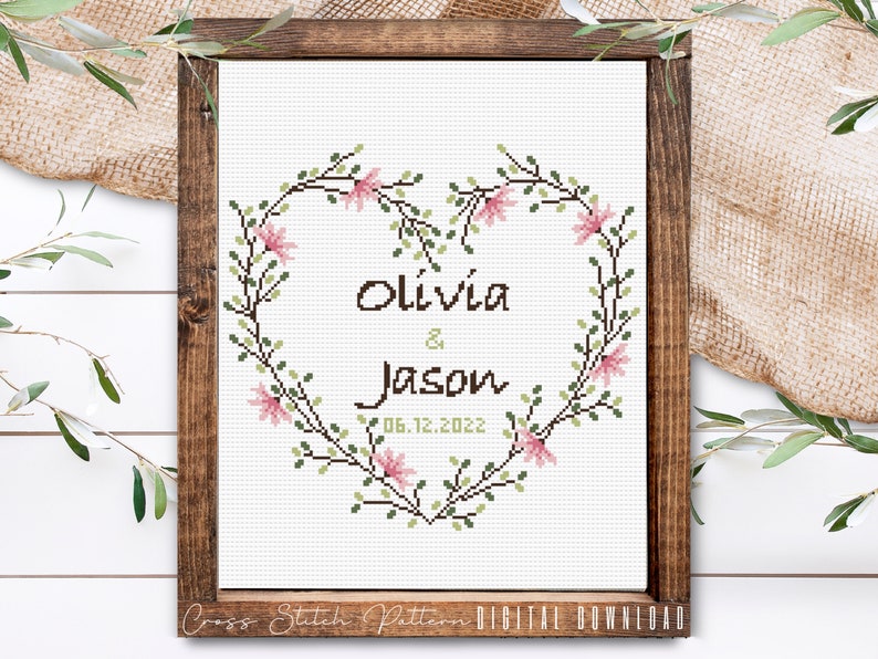 K&ouml;nnte beinhalten: Kreuzstichmuster mit einem herzf&ouml;rmigen Kranz aus gr&uuml;nen Bl&auml;ttern und rosa Bl&uuml;ten mit den Namen Olivia & Jason und dem Datum 06.12.2022.
