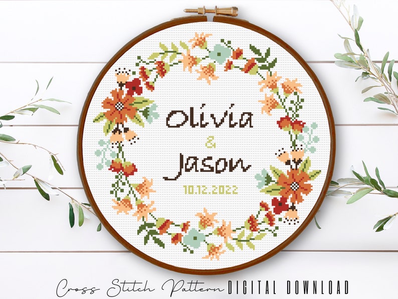 Puede incluir: Patr&oacute;n de punto de cruz con un dise&ntilde;o de corona de flores. La corona est&aacute; en tonos de naranja, rojo, verde y azul. El texto en el centro dice "Olivia & Jason 10.12.2022".