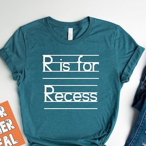 May include: A teal blue t-shirt with white text that reads "R is for Recess". The shirt is tied in a knot at the bottom. The t-shirt is laying on a white surface with a pair of blue jeans, a small wooden tray with a camera, and a small orange card that reads "The color is heather deep teal".