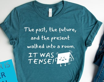 Op de afbeelding: Een turquoise T-shirt met de tekst "The past, the future, and the present walked into a room. It was tense!" in wit. Het shirt wordt getoond op een witte achtergrond met een blauwe jeans en witte sneakers.