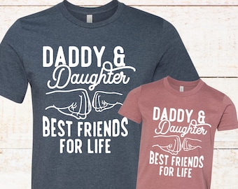 May include: Two t-shirts with a white graphic print of two fists bumping with the text "Daddy & Daughter Best Friends For Life". The larger shirt is navy blue and the smaller shirt is pink.