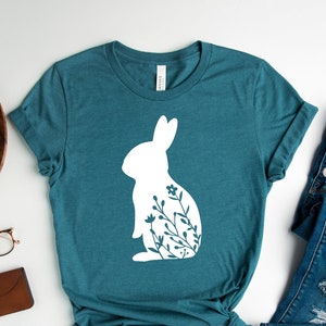 May include: Teal t-shirt with a white rabbit design filled with floral patterns. The image includes blue jeans and white trainers. A wooden tray with a plant, camera, watch, glasses, and wallet is also in the image.