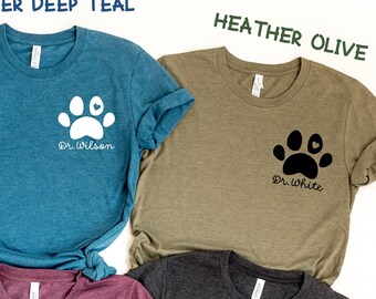 May include: A set of ten t-shirts in various colors, each featuring a white paw print with a heart in the center. The shirts have different text on them, including "Dr. Audrey", "Dr. Wilson", "Dr. White", "Vet Tech", "Future Vet", and "Amelia". The shirts are arranged in a circle on a white background.