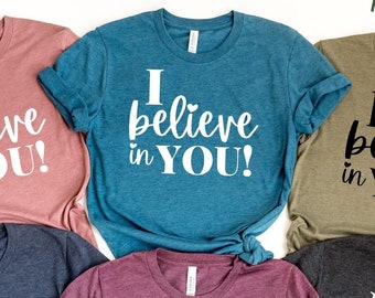 May include: Six t-shirts in different colors with the text "I believe in YOU!" printed in white on each shirt. The shirts are laid out on a white background.