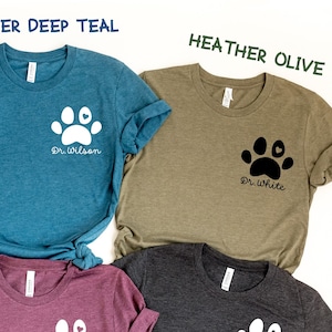 May include: A set of ten t-shirts in various colors, each featuring a white paw print with a heart in the center. The shirts have different text on them, including "Dr. Audrey", "Dr. Wilson", "Dr. White", "Vet Tech", "Future Vet", and "Amelia". The shirts are arranged in a circle on a white background.