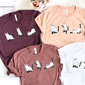 May include: Five t-shirts with a graphic of cats dressed as ghosts. The shirts are in various colors, including maroon, peach, black, brown, and white.