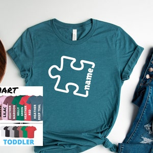 May include: A teal t-shirt with a white puzzle piece design and the text "name" on the front. The shirt is available in a variety of colors and sizes.