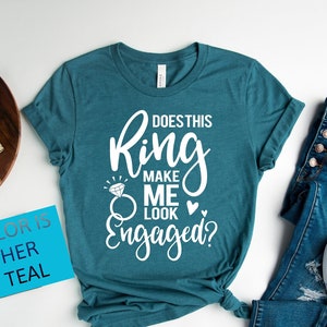 May include: A teal t-shirt with the text "Does this ring make me look engaged?" in white. The shirt is tied in a knot at the bottom. The color of the shirt is Heather Deep Teal.