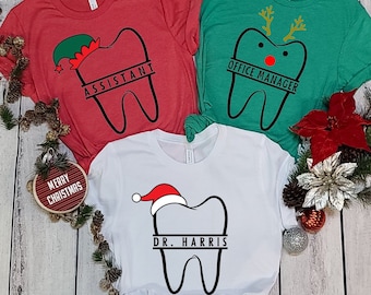 May include: Three Christmas-themed t-shirts featuring a tooth design. The red shirt says "Assistant" with an elf hat on the tooth. The green shirt says "Office Manager" with reindeer antlers on the tooth. The white shirt says "Dr. Harris" with a Santa hat on the tooth.