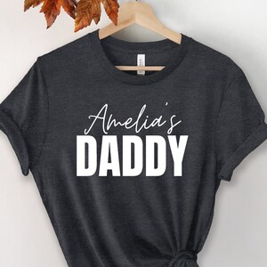 May include: Dark grey t-shirt with white text that reads "Amelia's Daddy".