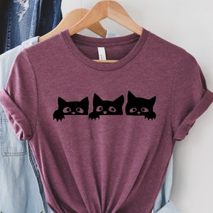 May include: A maroon t-shirt with a graphic of three black cats peeking over a surface. The cats have white eyes and are looking at the viewer.