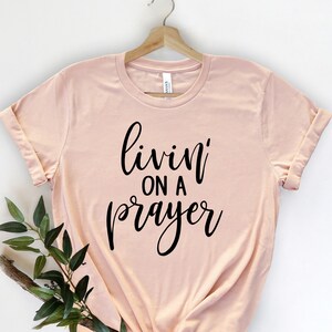 Peut inclure: Un t-shirt rose p&acirc;le avec le texte "livin' on a prayer" en &eacute;criture noire.