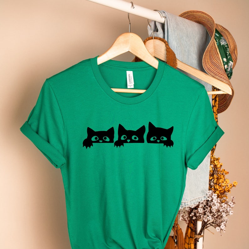 May include: A green t-shirt with a black graphic of three cats peeking over a surface. The cats are all facing the same direction and have the same expression.