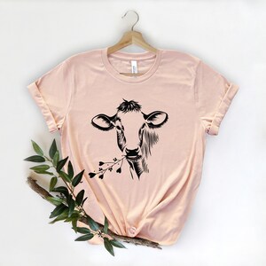 May include: A peach-colored t-shirt with a black silhouette of a cow eating a sprig of flowers. The t-shirt is hanging on a wooden hanger.