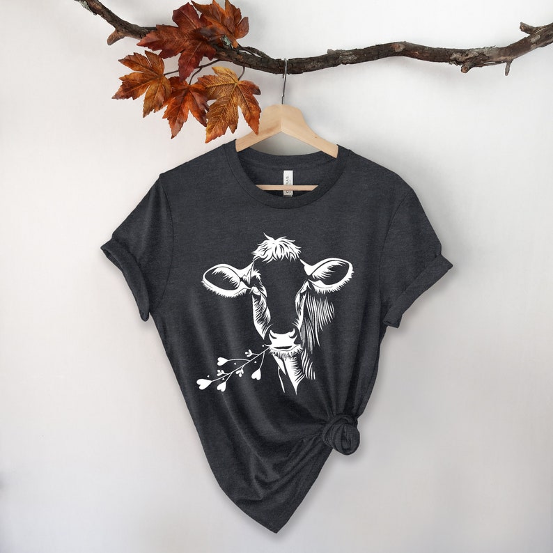 May include: A dark heather grey t-shirt with a white silhouette of a cow eating a sprig of flowers.