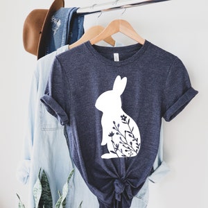 May include: A navy blue t-shirt with a white rabbit silhouette and floral details. The t-shirt is displayed on a wooden hanger alongside a light blue shirt and a brown hat, suggesting a casual clothing theme.