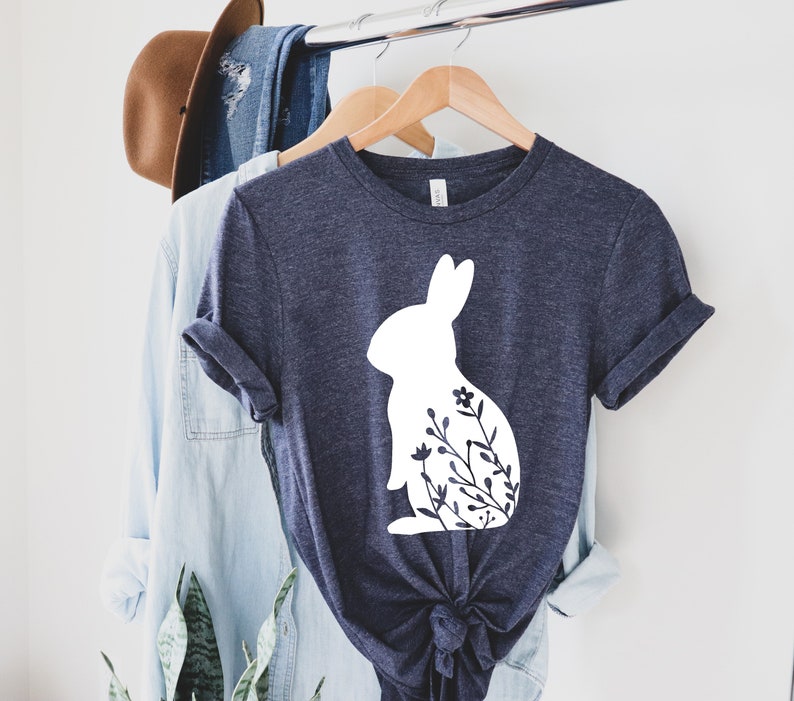 May include: A navy blue t-shirt with a white rabbit silhouette and floral details. The t-shirt is displayed on a wooden hanger alongside a light blue shirt and a brown hat, suggesting a casual clothing theme.