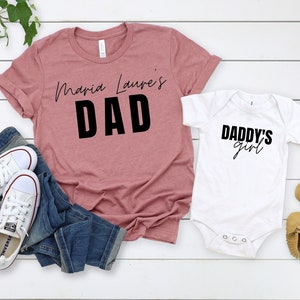 May include: A set of two shirts, one pink and one white. The pink shirt says "Maria Laura's Dad" in black text. The white shirt says "Daddy's Girl" in black text.