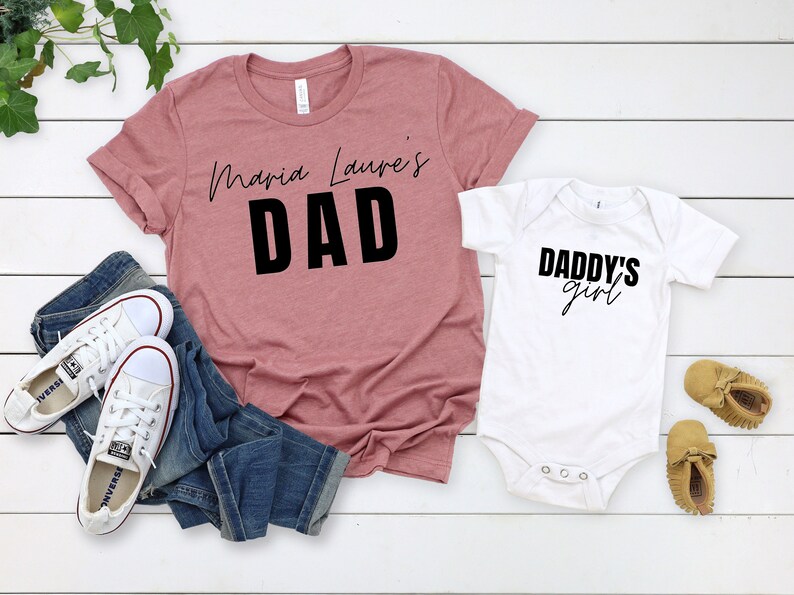 May include: A set of two shirts, one pink and one white. The pink shirt says "Maria Laura's Dad" in black text. The white shirt says "Daddy's Girl" in black text.